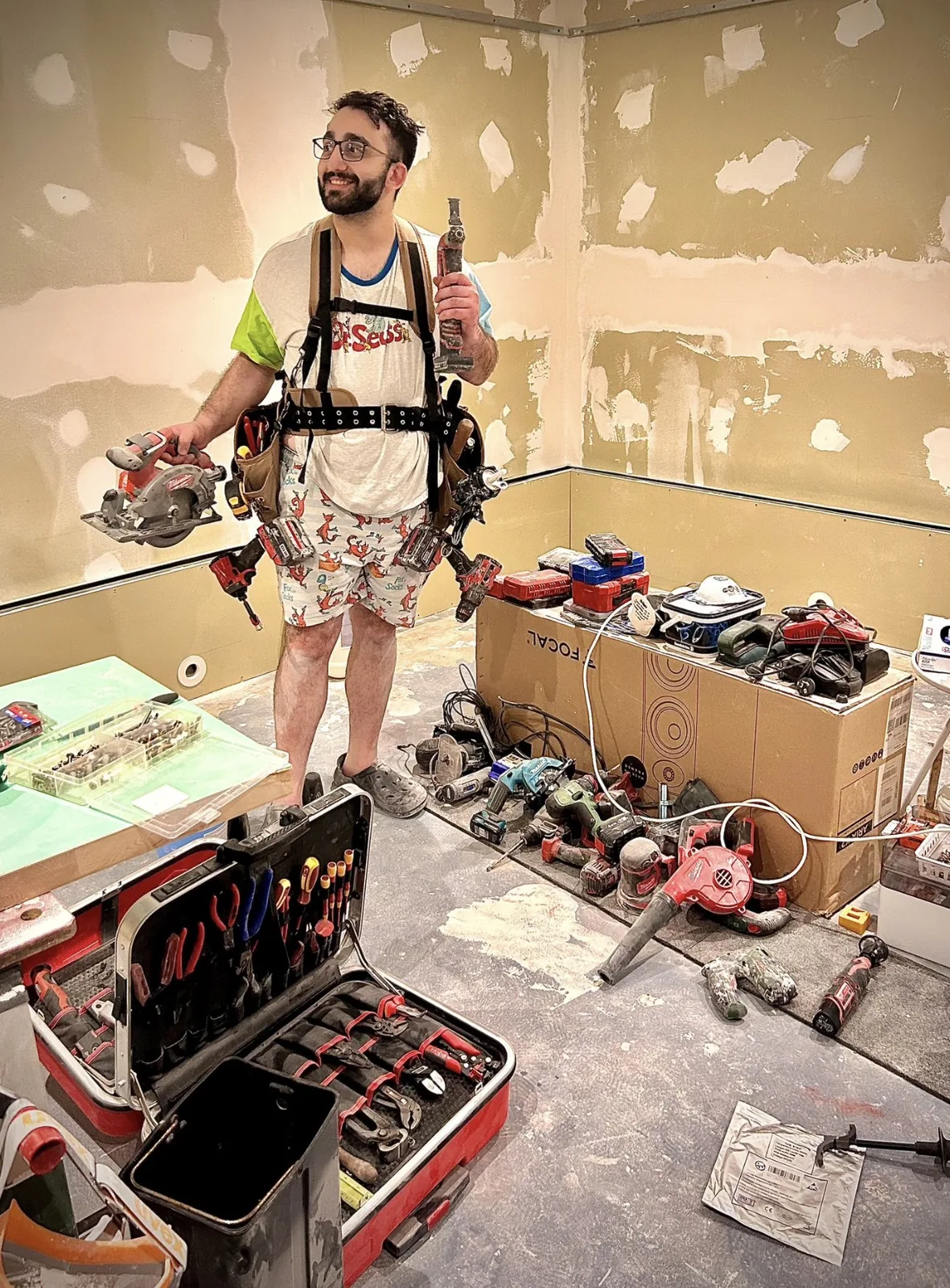 Arman in a tool harness and pyjama shorts, surrounded by power tools.