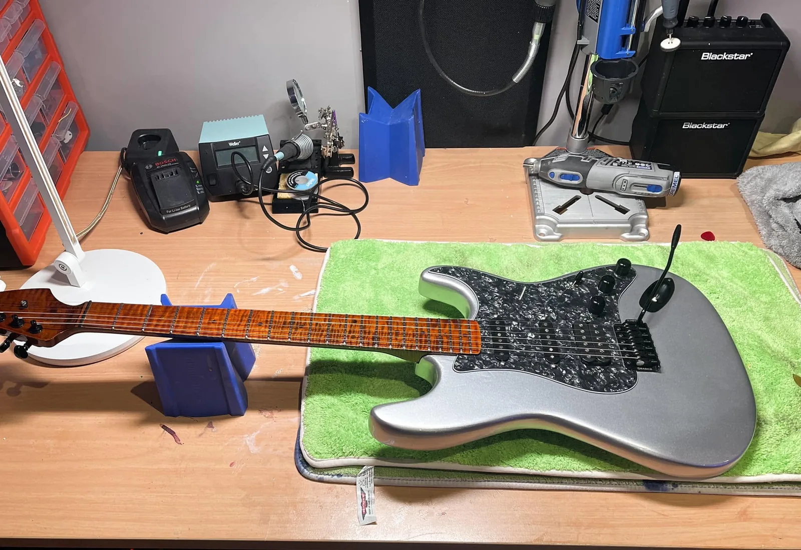 A silver guitar on a workbench beside a soldering station.